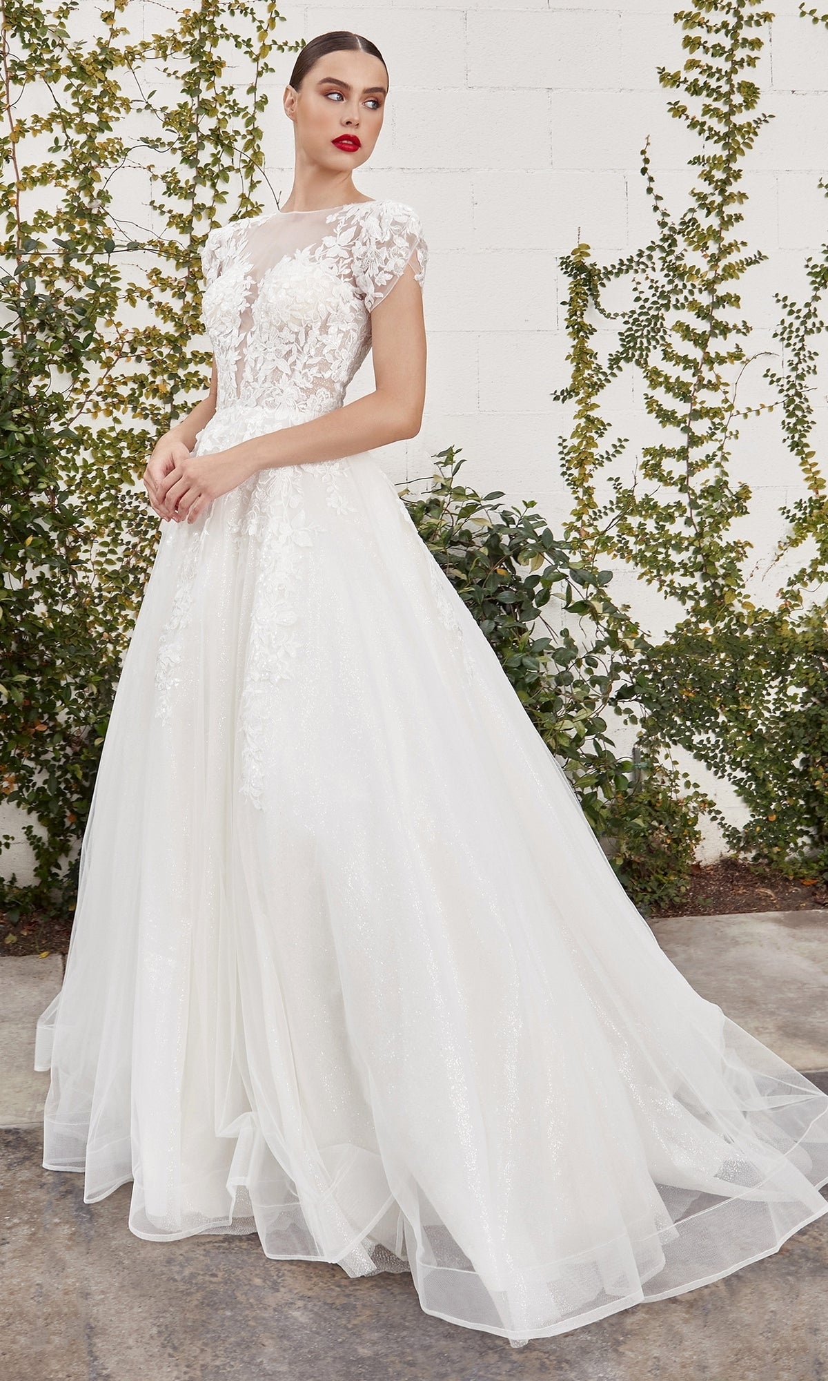 A woman in the Cap-Sleeve White Wedding Gown: Ladivine A1082W, featuring floral lace and a full tulle skirt, stands outdoors against a white brick wall and lush green foliage.